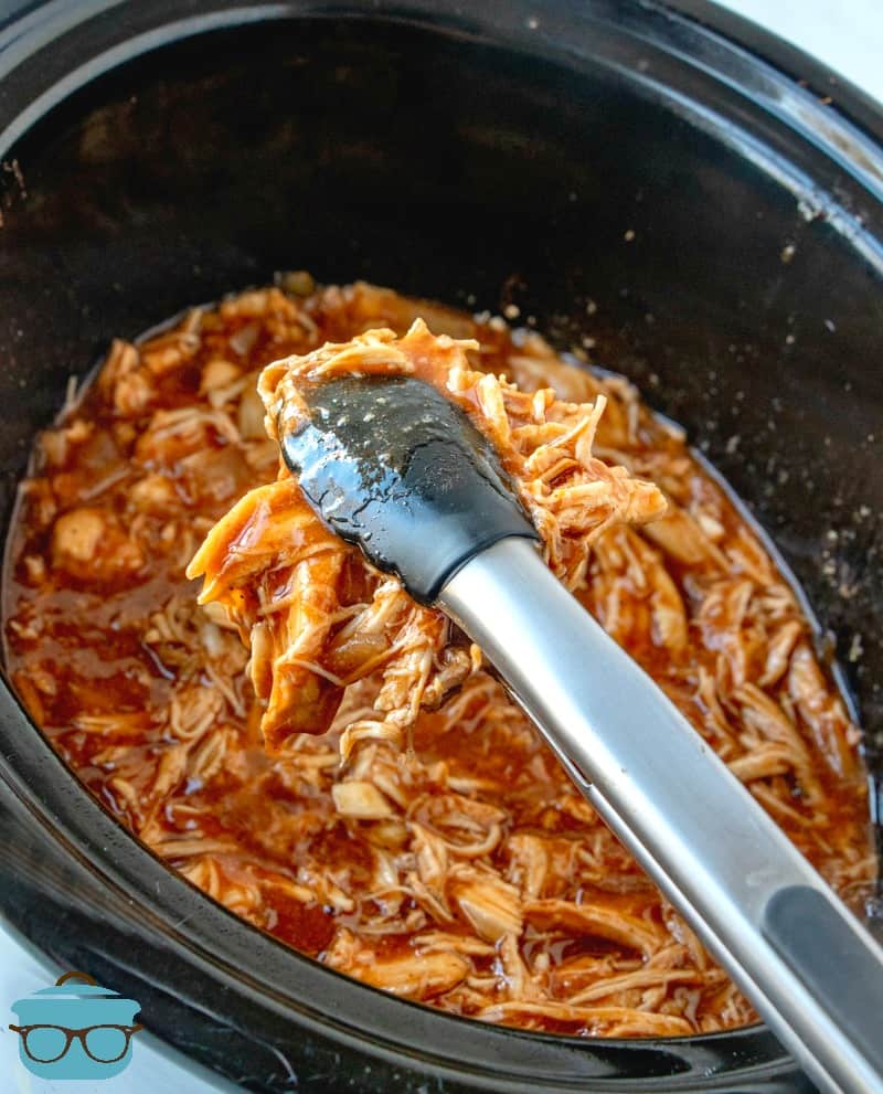 tongs scooping up shredded barbecue chicken out of a slow cooker.
