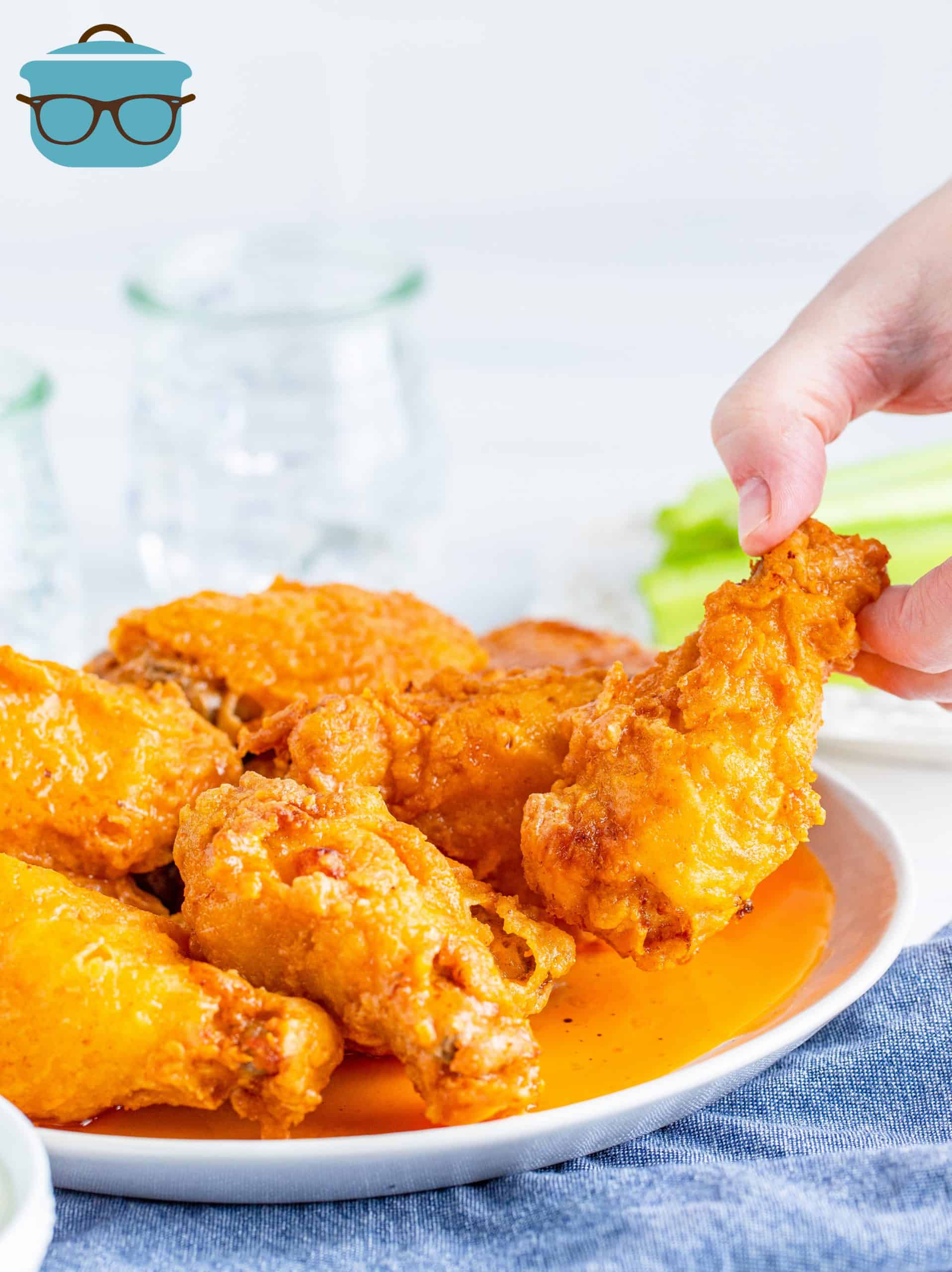 The Best Buffalo Chicken Wings shown on a white plate with one being removed by a person's hand to the side.