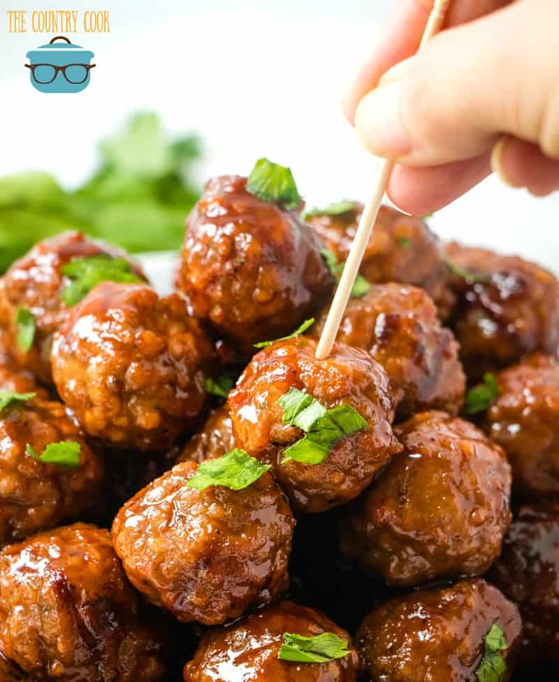 a hand pushing a toothpick into a meatball that is at the top of a pile of other meatballs.