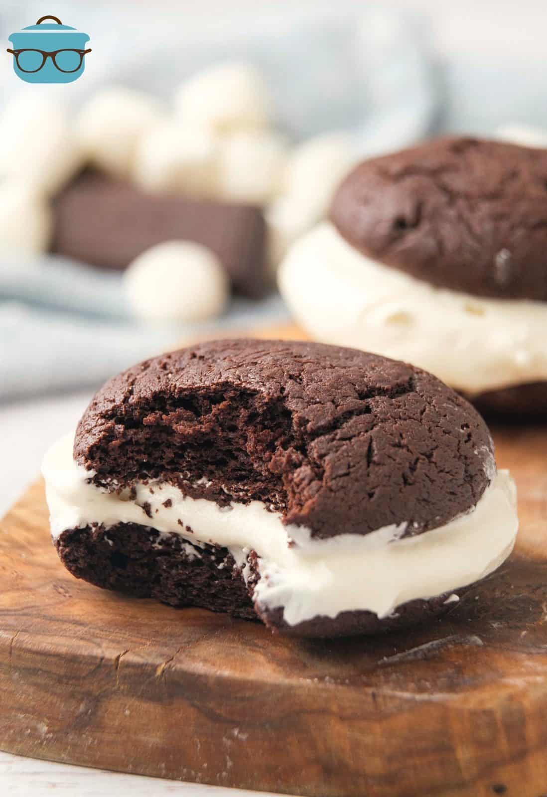 a chocolate whoopie pie with a bite removed is sitting on a wooden cutting board.