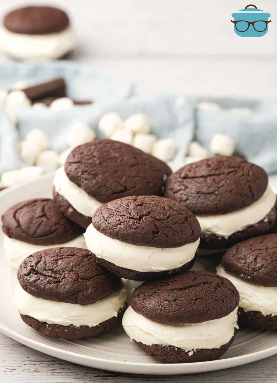 A stack of whoopie pies on a round white plate with marshmallows and pieces of chocolate in the background.
