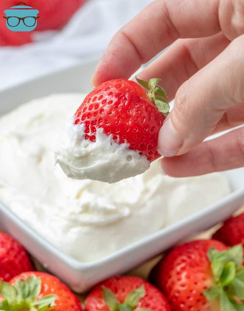 pictured: hand holding a whole strawberry that has been dipped in white fruit dip surround by additional strawberries.