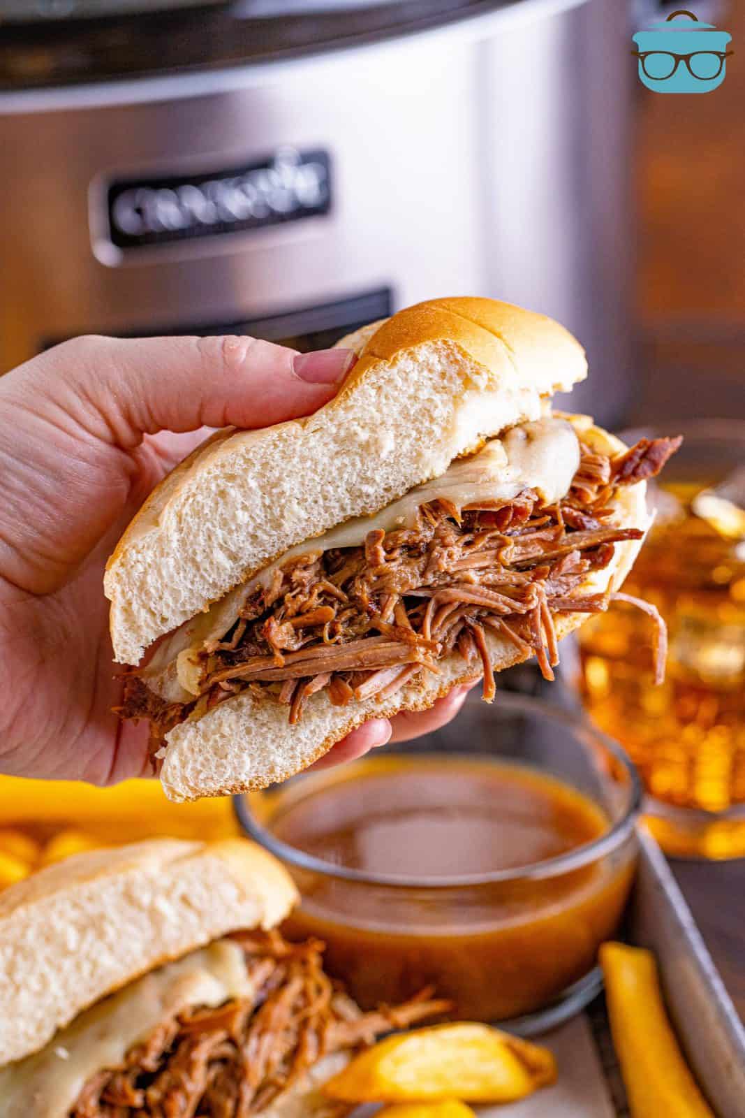 hand holding a slice of beef dip sandwich in front of a slow cooker. 