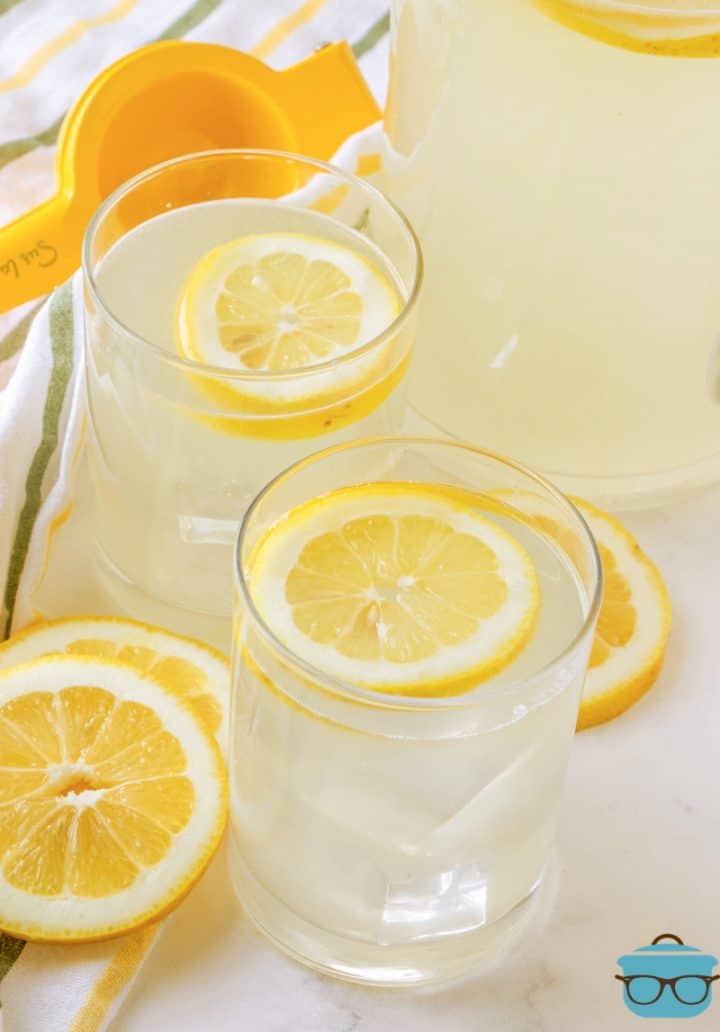 freshly squeezed lemonade shown served in two glass topped with lemon slices and a pitcher of lemonade in the background