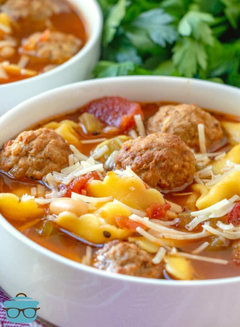 close-up Meatball and Tortellini soup in a white bowl topped with shredded Parmesan cheese with a cilantro bunch in the background.