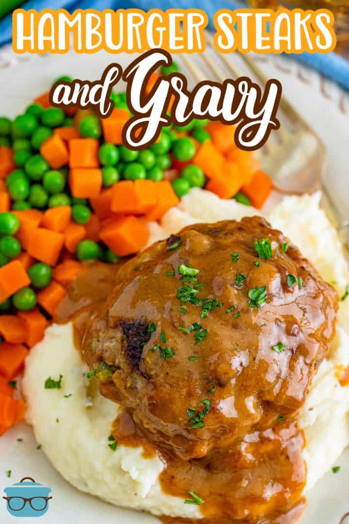 a hamburger patty covered in gravy on a bed of mashed potatoes and peas and carrots on the side.
