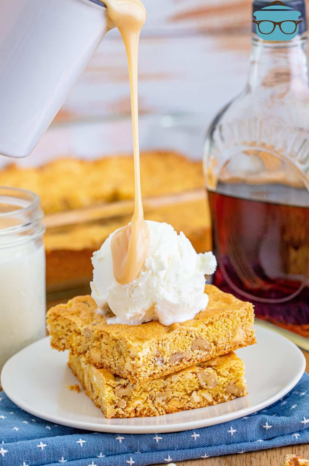 maple butter sauce being shown pouring on top of two slices of blondies on a white plate.
