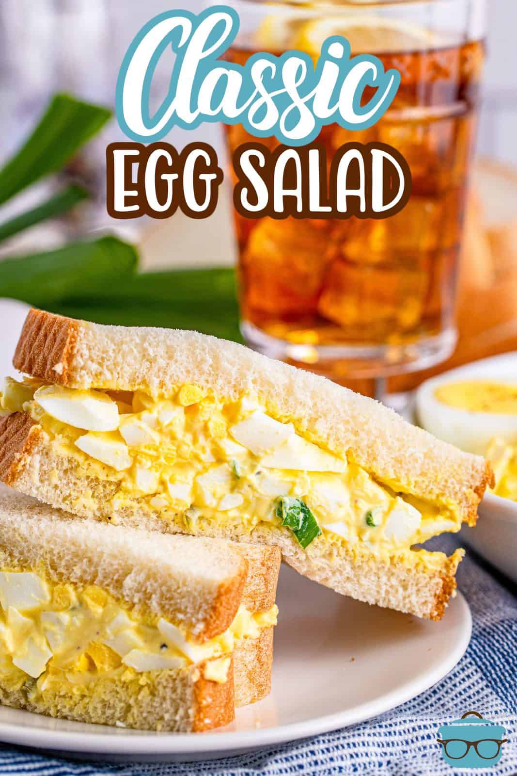 two half egg salad sandwiches stacked on top of each other with a glass of iced tea in the background.