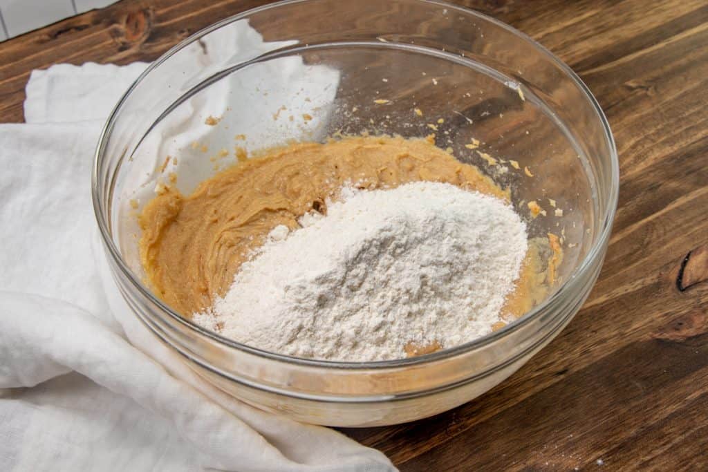 adding flour mixture to peanut butter mixture in a large bowl on a wooden surface