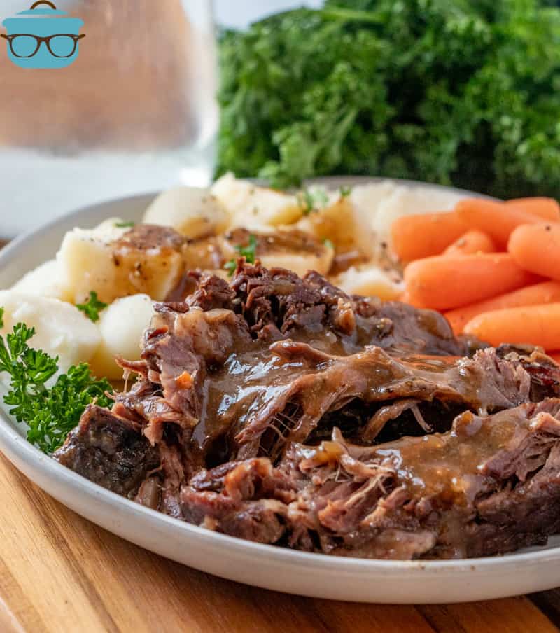 Crock Pot Pot Roast, slices served on a round white plate and served with baby carrots and diced potatoes