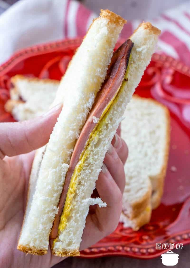 a hand holding up a sliced, fried bologna with yellow mustard and white bread