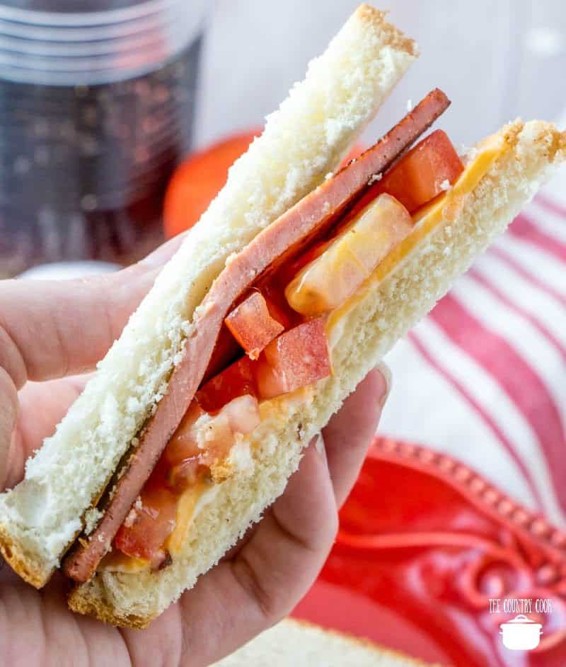 a hand holding one slice of fried bologna sandwich that has tomatoes and cheese on it.