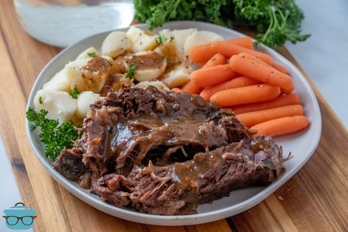 Slow Cooker Pot Roast on a white plate and topped with gravy served with carrots and potatoes with parsley in the background.