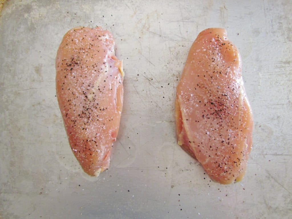 two chicken breasts on a baking tray covered in oil, salt and pepper