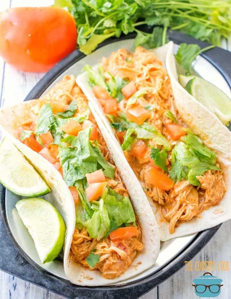 two shredded chicken tacos on a plate with two fresh sliced limes on the side. 