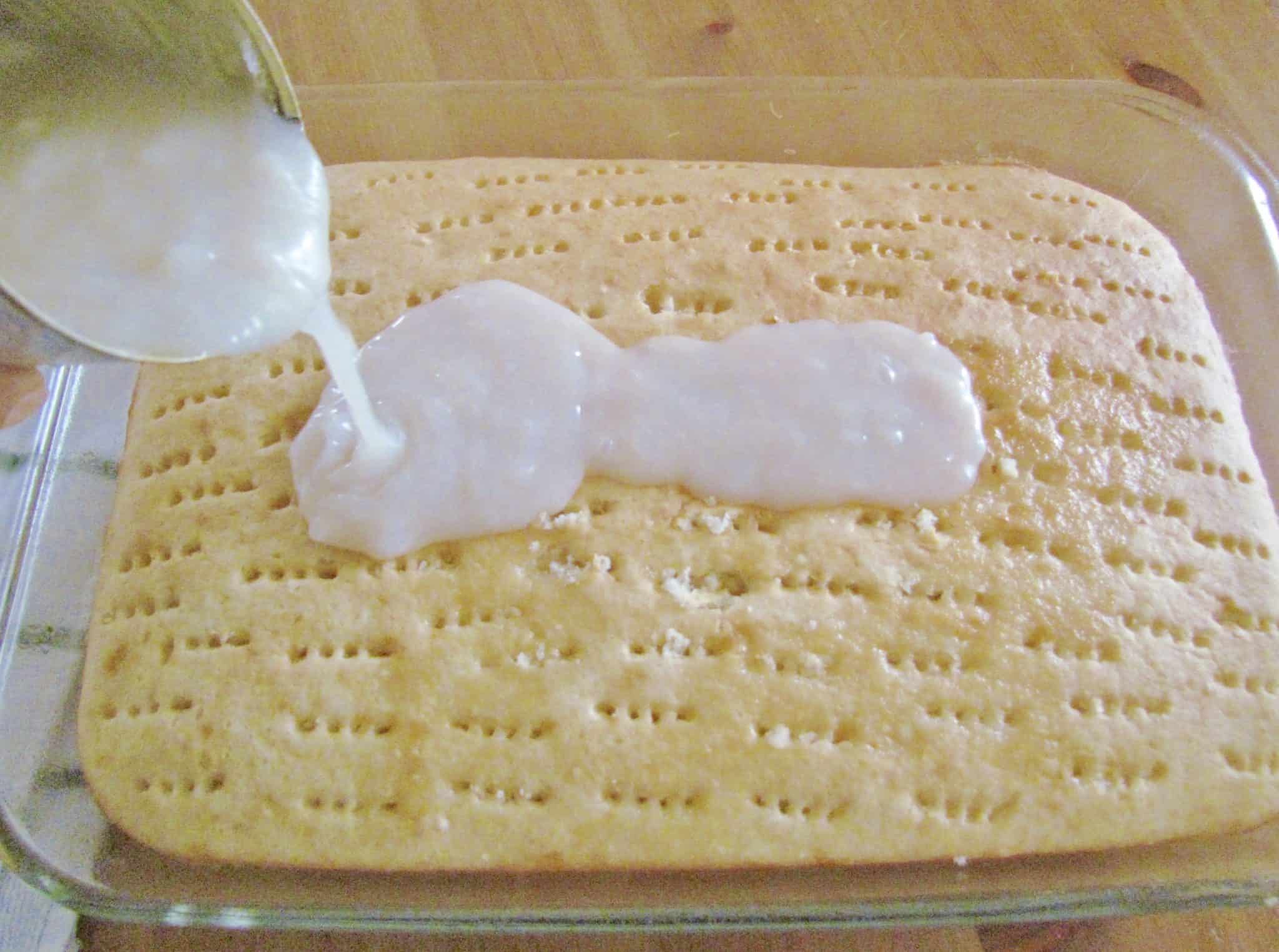 pouring cream of coconut into the poked holes of baked white cake.