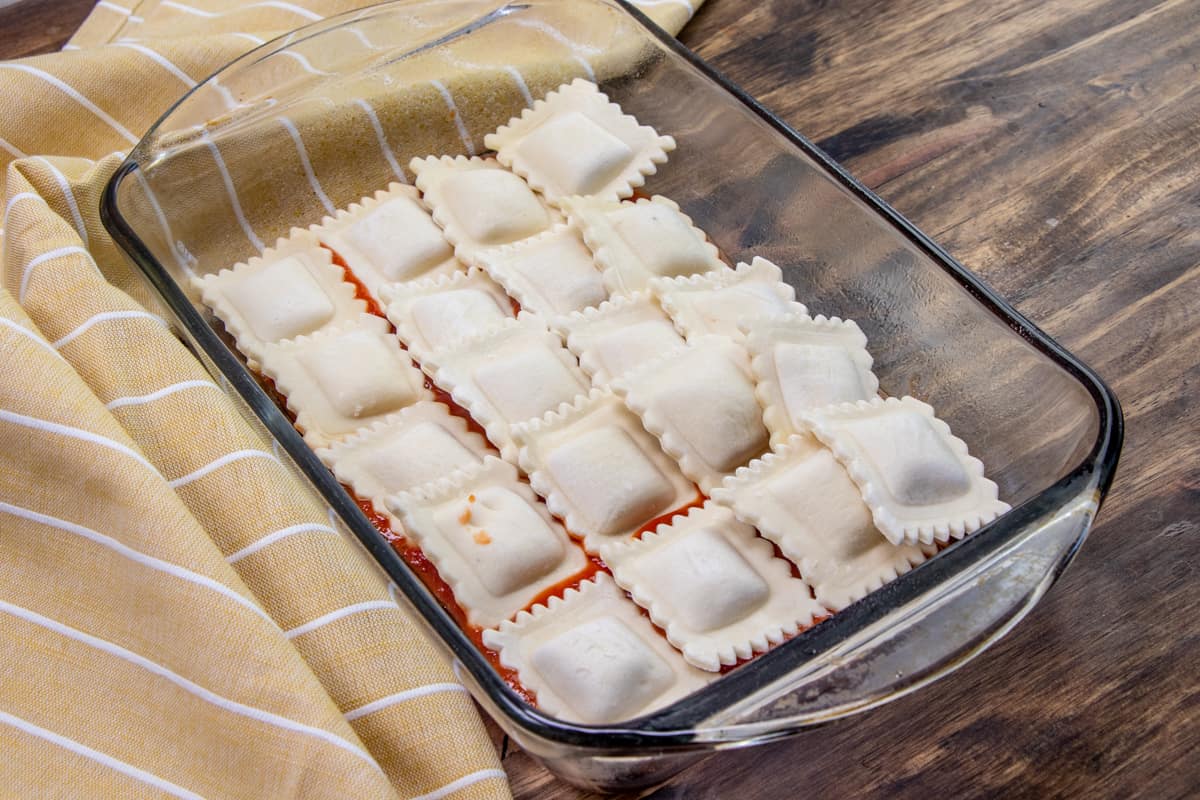 a layer of pasta sauce on the bottom of a clear 11-inch by 7-inch baking dish topped with a layer of frozen ravioli.