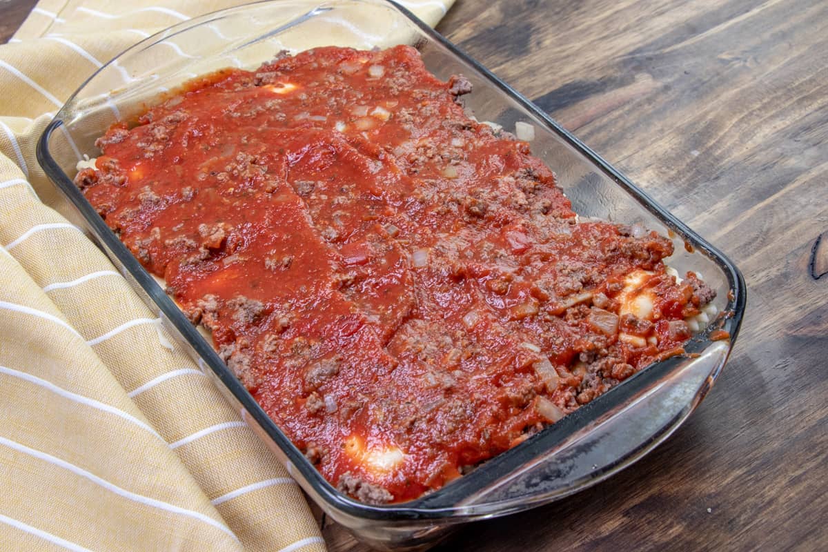 a layer of pasta sauce on top of cooked ground beef and frozen ravioli in a Pyrex glass baking dish.