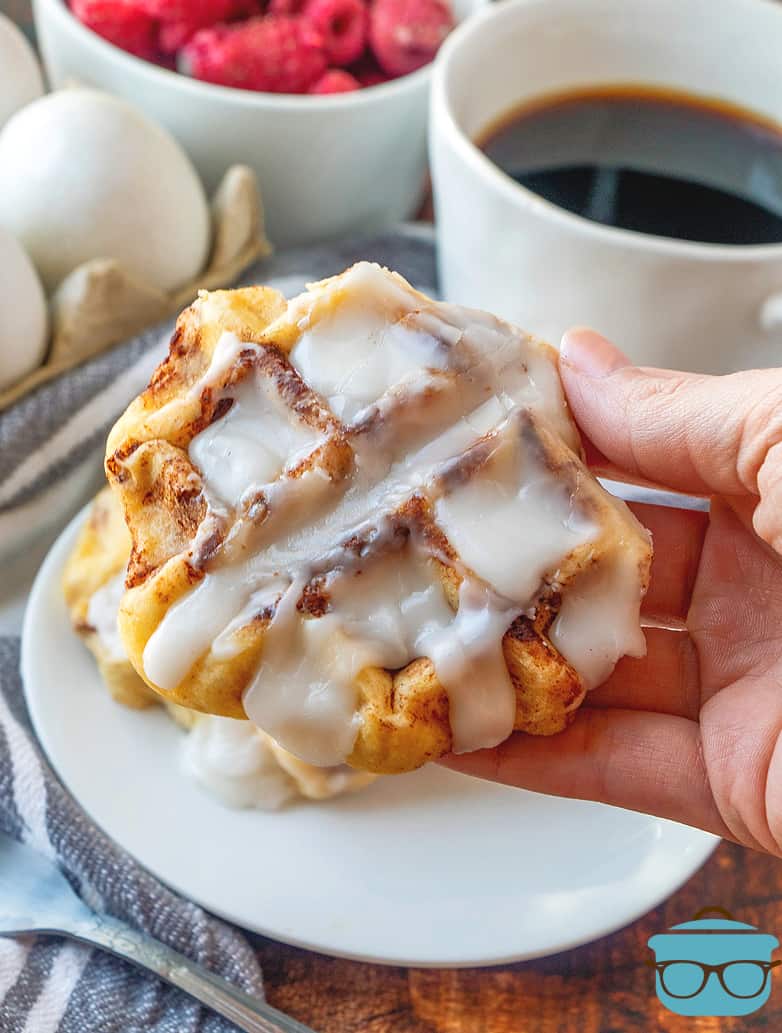 a cinnamon roll waffle being held in a hand with a cup of coffee in the background.