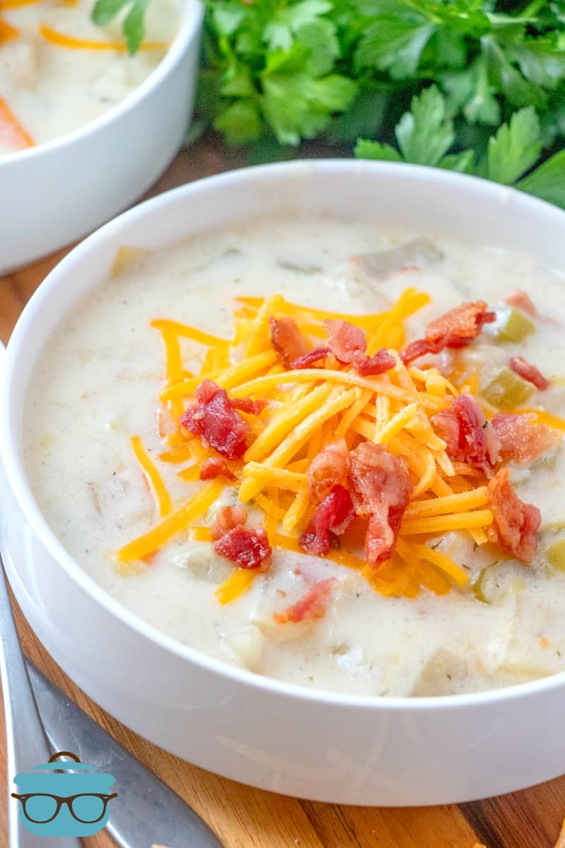Potato Leek Soup shown in a white bowl, topped with shredded cheddar cheese and crumbled bacon, parsley pictured in the background.