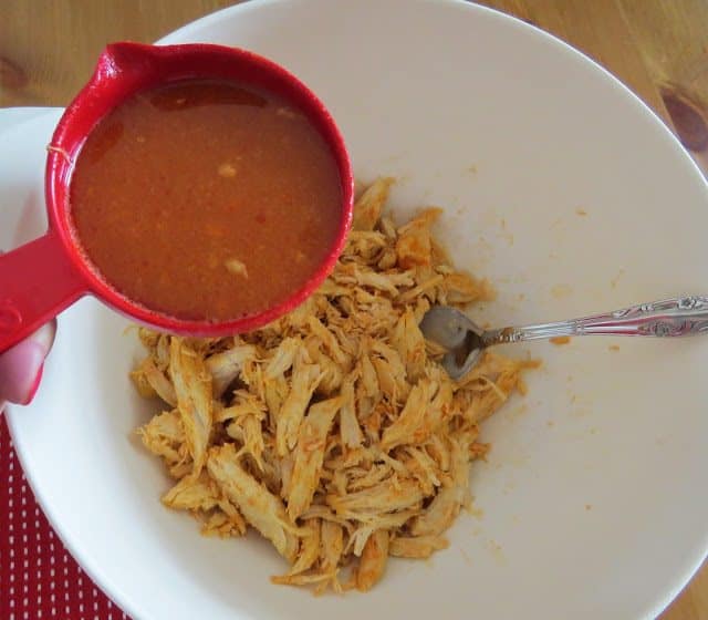 pouring buffalo chicken cooking liquid over shredded cooked chicken in a bowl