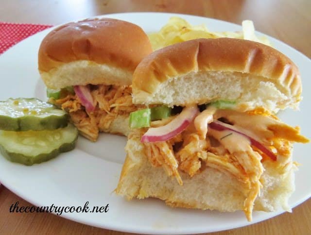 two smalll buffalo chicken sliders with celery slaw on a white plate with two sliced pickles and potato chips.