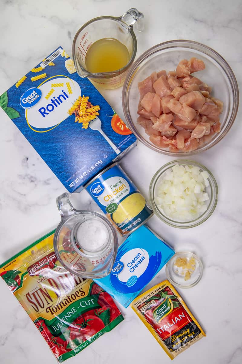 ingredients for creamy Italian chicken: boneless, skinless chicken breasts, chicken broth, Italian salad dressing mix, chopped onion, minced garlic, cream of chicken soup, cream cheese, rotini pasta, sun-dried tomatoes.