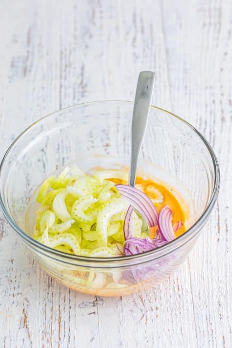 A glass mixing bowl with red onion, buffalo sauce, and celery.