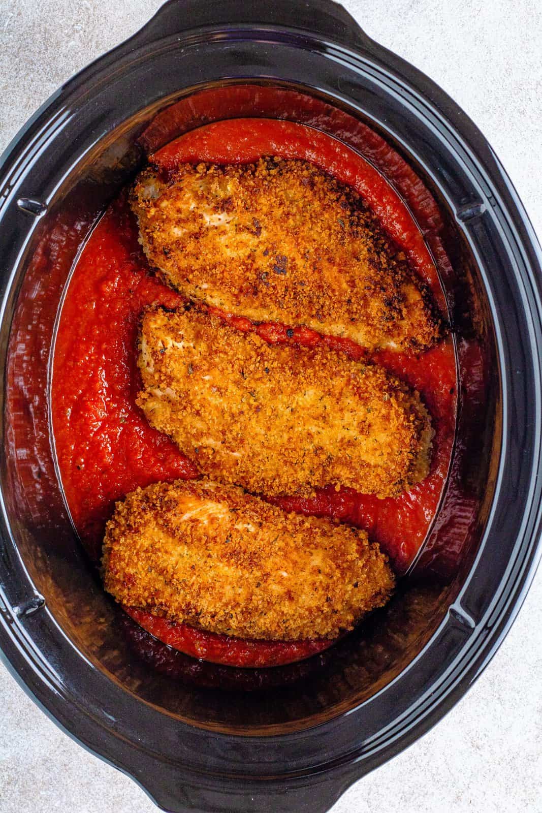 browned chicken breasts shown on top of sauce in an oval slow cooker.