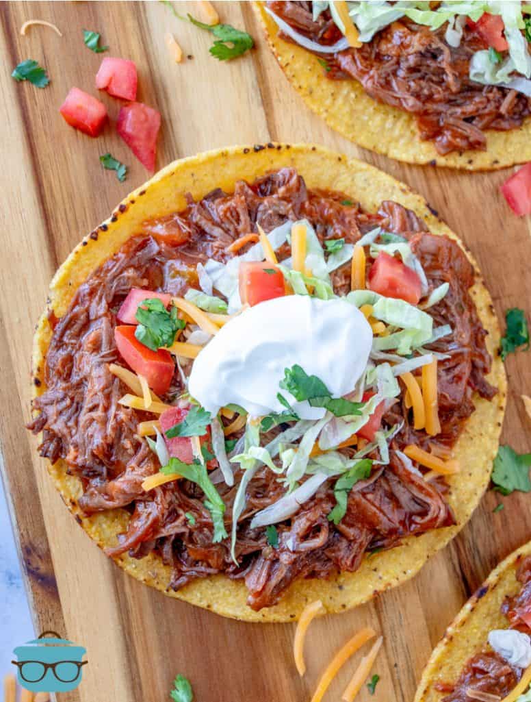 tostada with shredded beef, lettuce and tomato on a wooden cutting board.