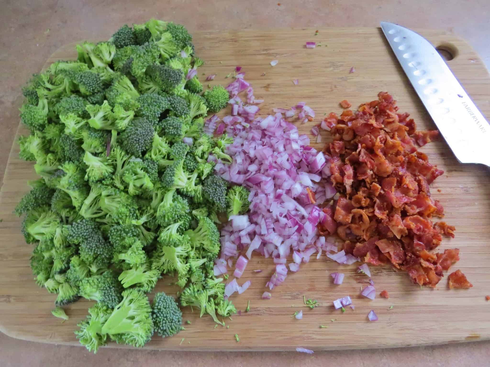 chopped broccoli, diced red onion, crumbled and cooked bacon on a wooden chopping board with knife.