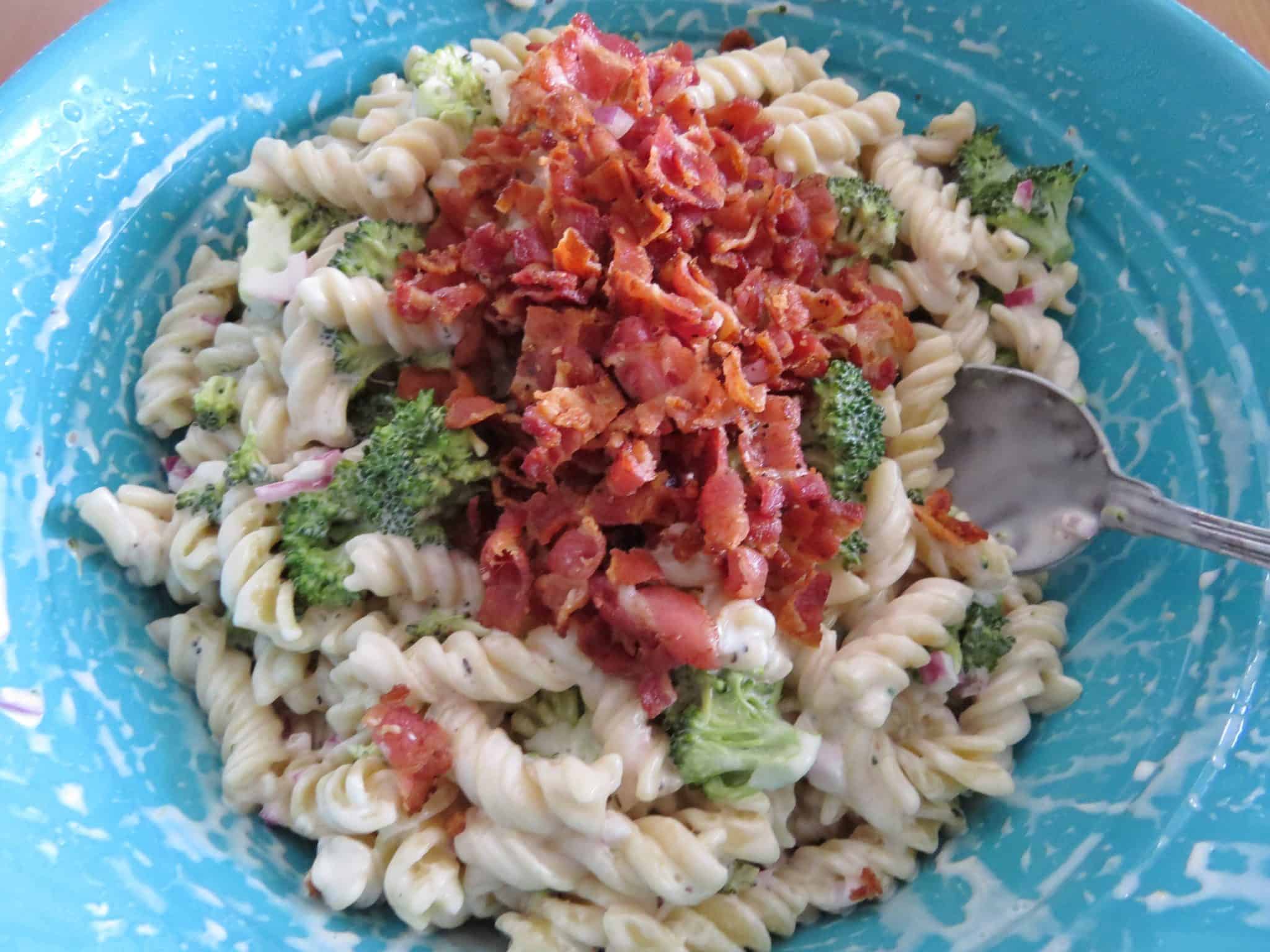 cooked crumbled bacon stirred together with broccoli florets, diced red onion and cooked rotini pasta and creamy dressing in a blue bowl.