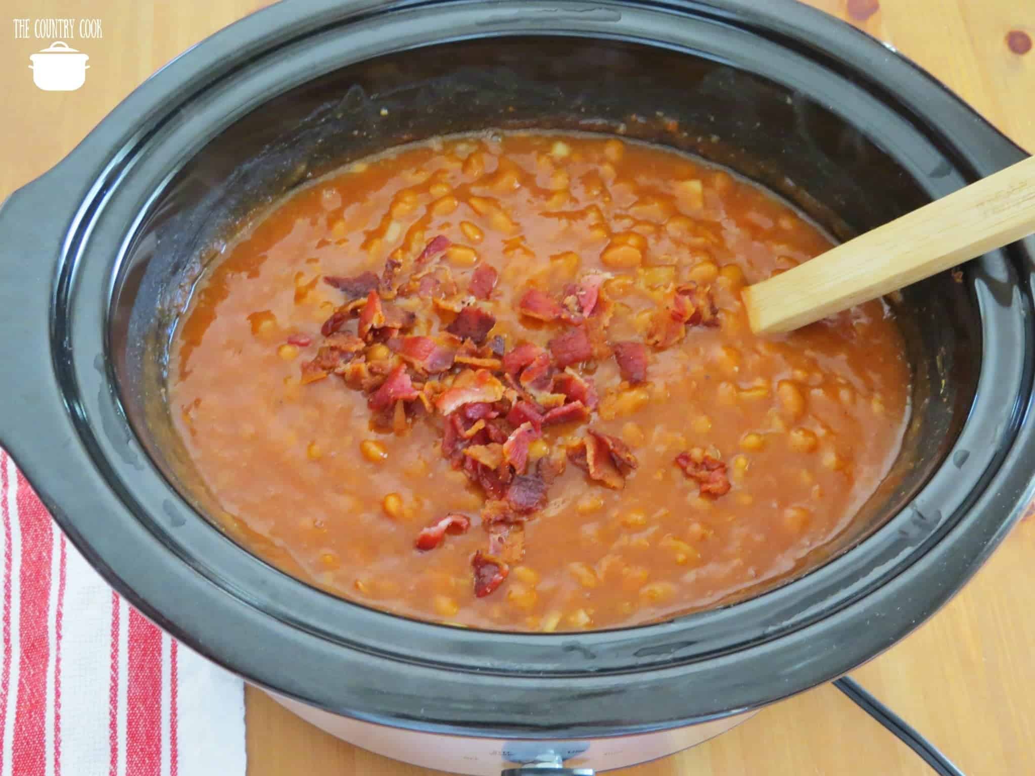 chopped cooked bacon shown on top of cooked beaked beans in crock pot. 