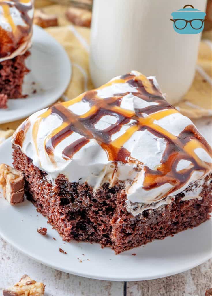 chocolate cake with Snickers whipped cream topping drizzled with chocolate and caramel, slice shown on a small white plate with a bottle of milk in the background