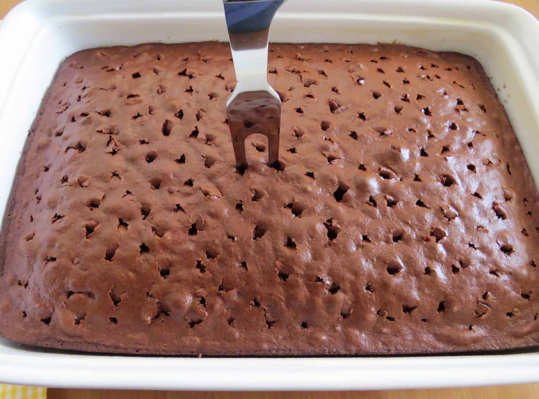 a large meat fork piercing holes in fully baked chocolate cake in a white baking dish.