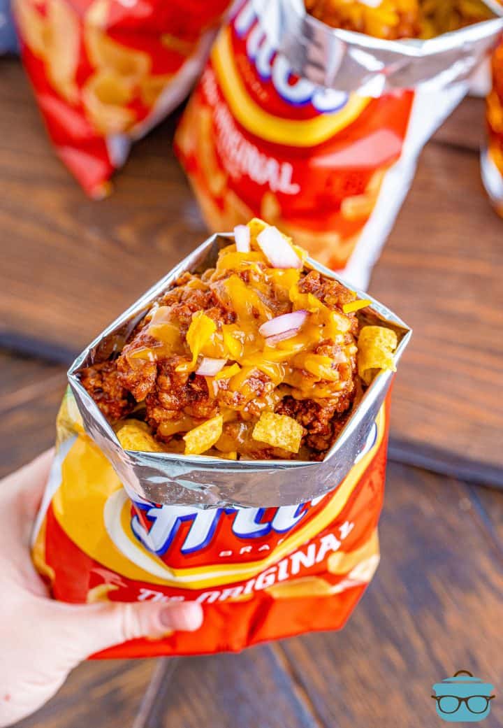 a hand holding a bag of Fritos corn chips that is topped with chili, cheese and diced onions.