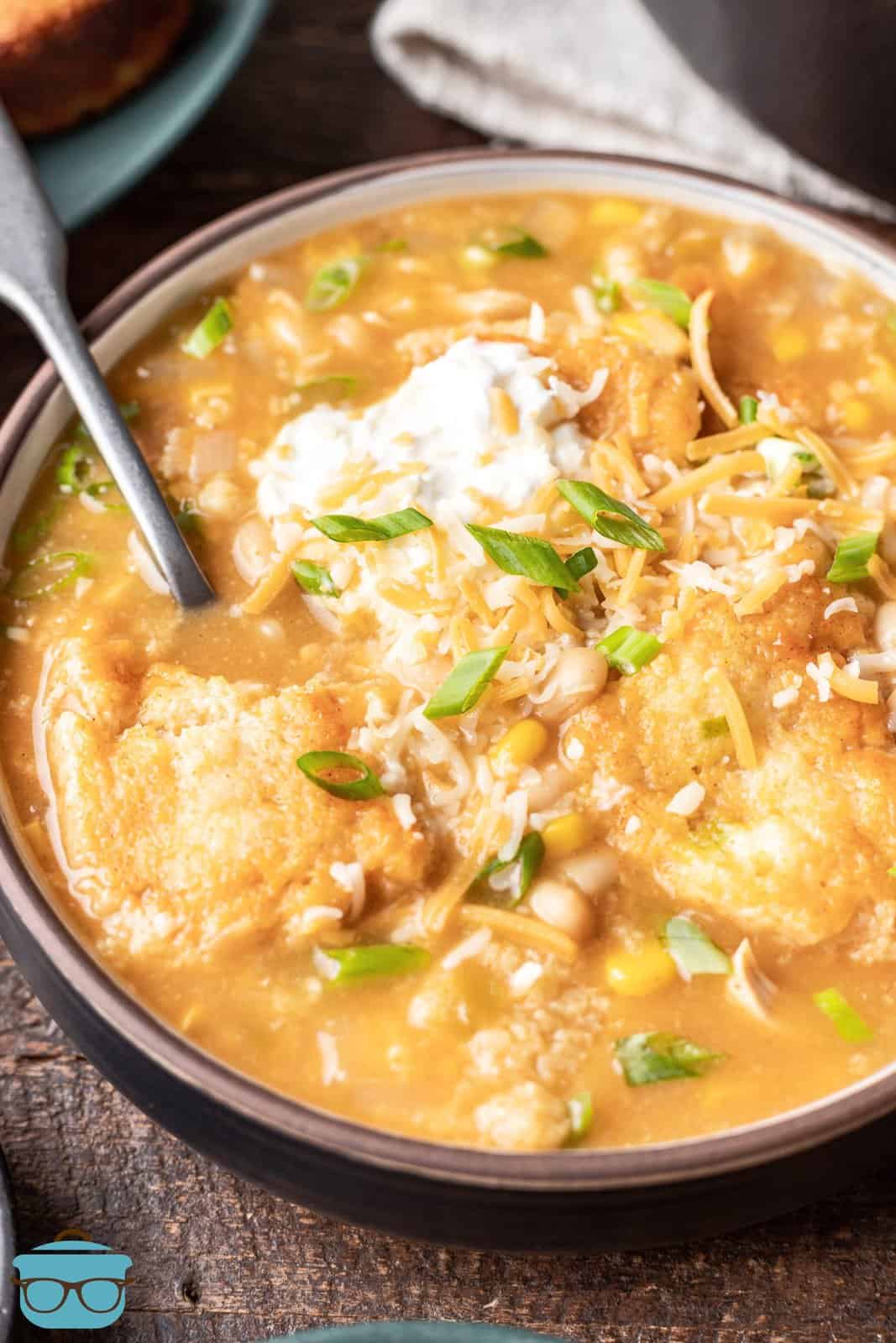 a closeup photo of a bowl of white chicken chili with cornbread dumplings with a spoon inserted and topped with a dollop of sour cream. 