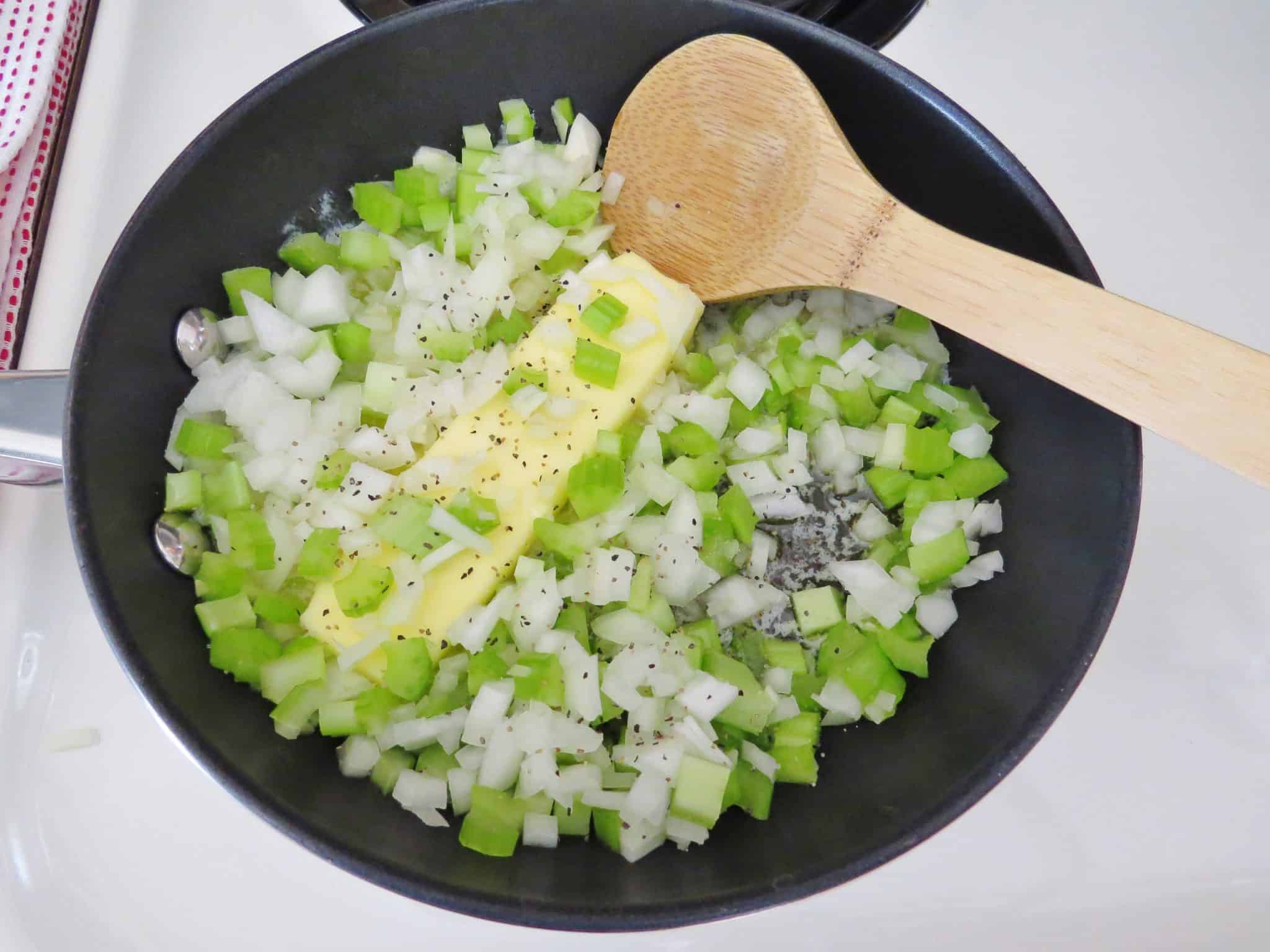 diced celery and diced onion with a stick of butter in a large skillet with a wooden spoon.