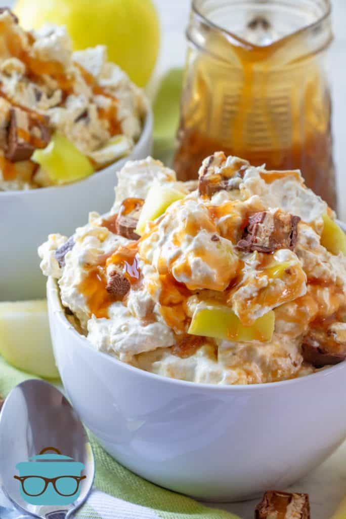 SNICKERS CARAMEL APPLE SALAD SHOWN IN A WHITE BOWL WITH A JAR OF CARAMEL SAUCE AND A SPOON