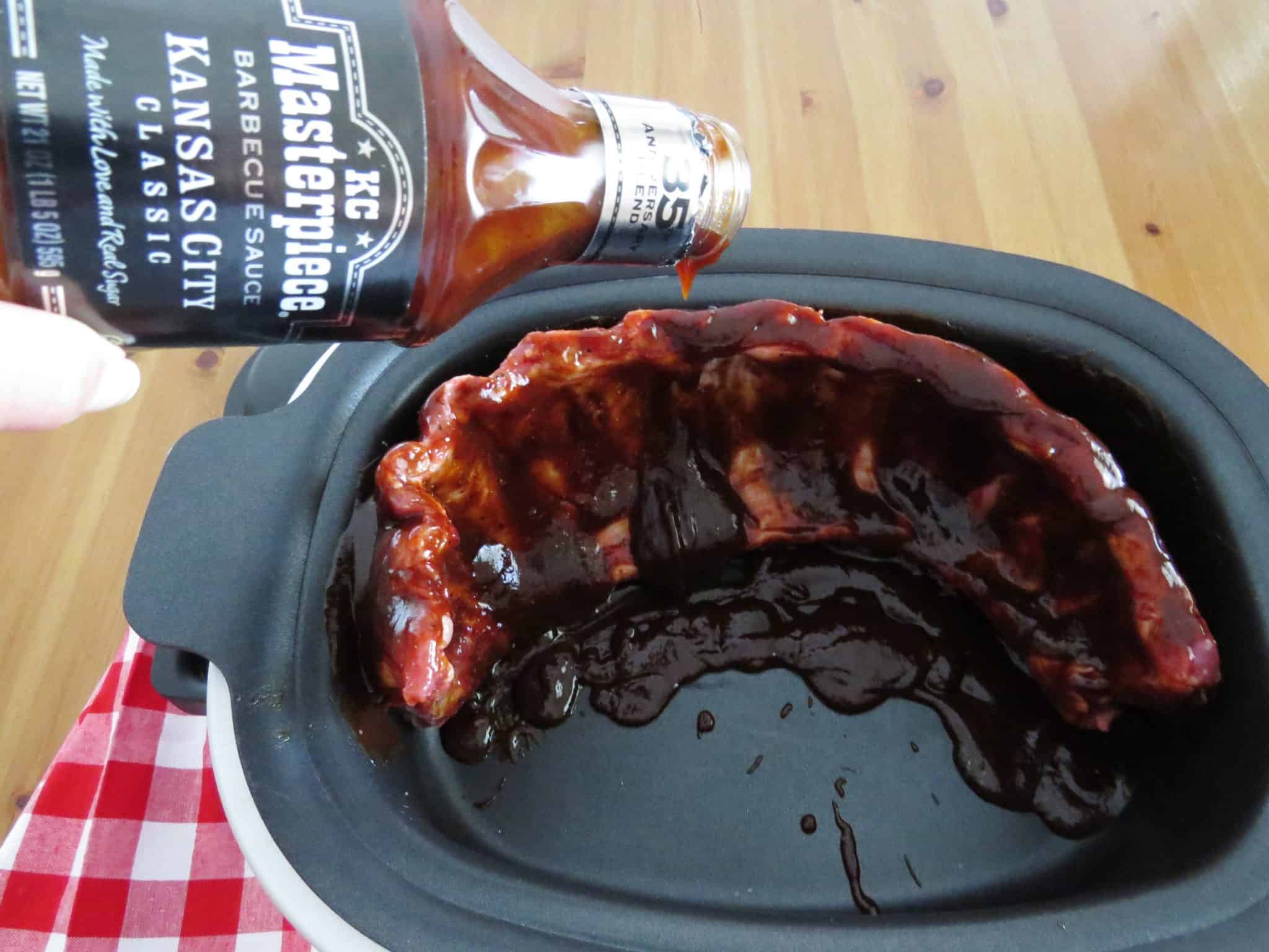 pouring a bottle of barbecue sauce over a rack of ribs inside an oval slow cooker.