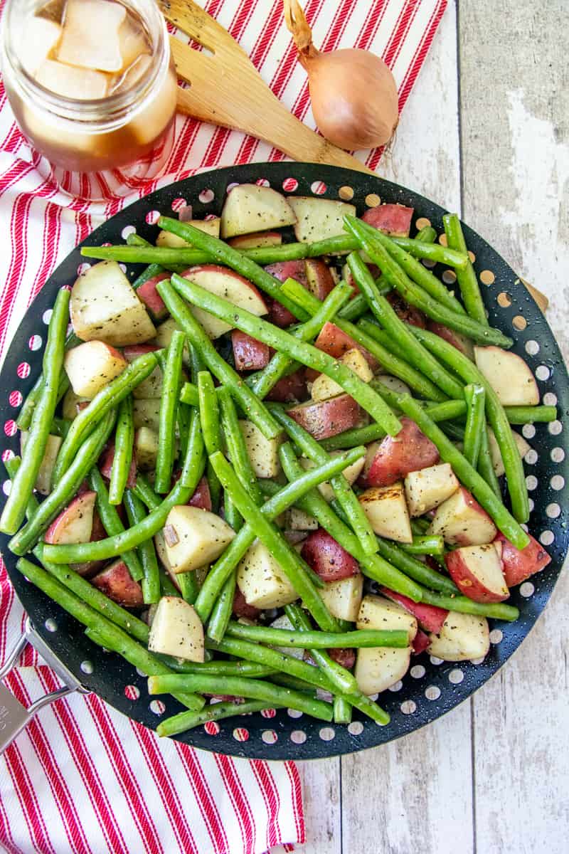 Diced potatoes and fresh green beans shown in a black grill pan with a glass of iced tea off to the side of the basket.