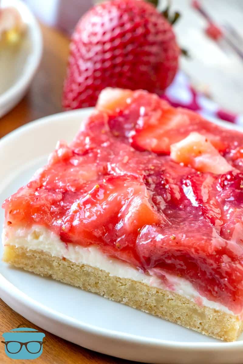 slice of Strawberries and Cream Cookie Bars on a white plate with a strawberry in the background.