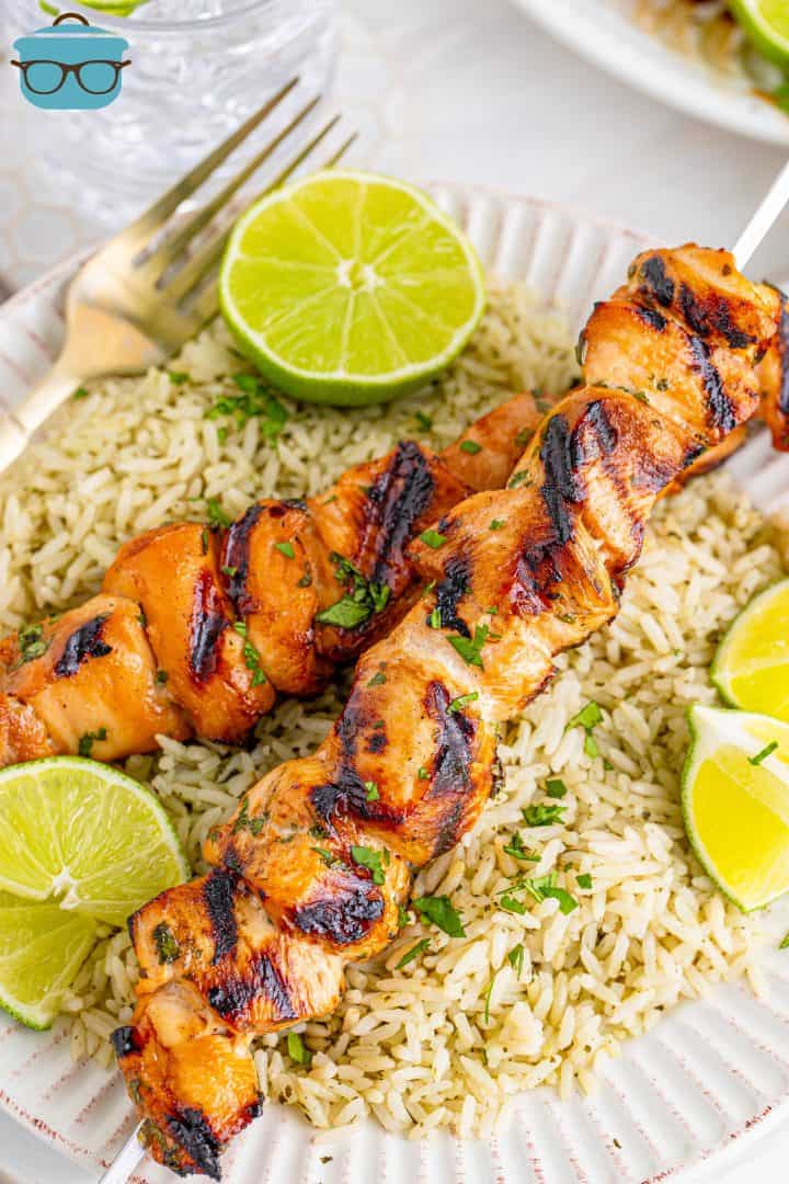 two grilled chicken skewers served over cilantro lime rice on a white plate with sliced fresh limes on the side.