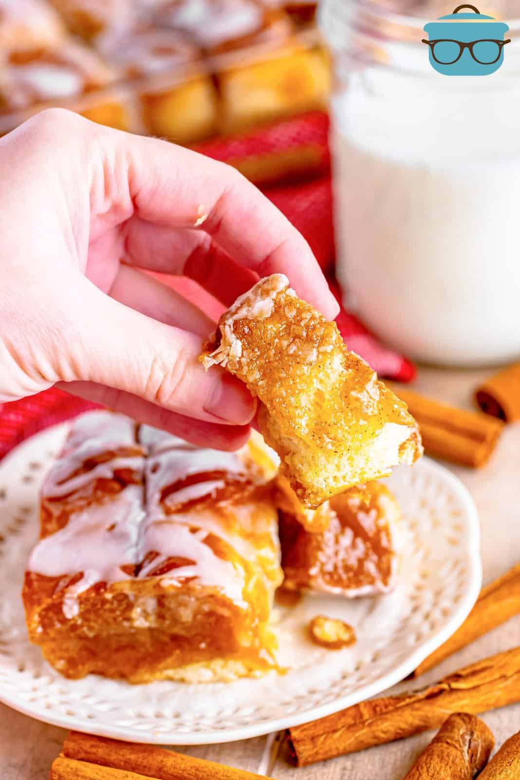 a hand holding up a bite of cinnamon pull apart in front of a plate and a glass of milk. 