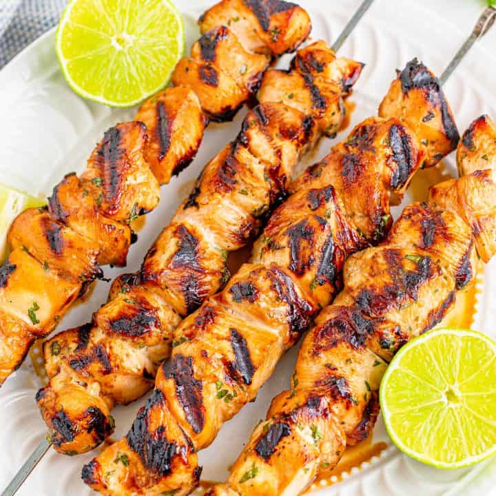 four grilled honey lime chicken skewers on a white plate with sliced fresh limes.