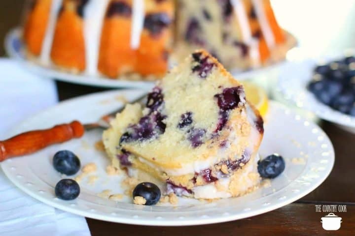 a slice of Blueberry Muffin Cake on a white plate surrounded by fresh blueberries.