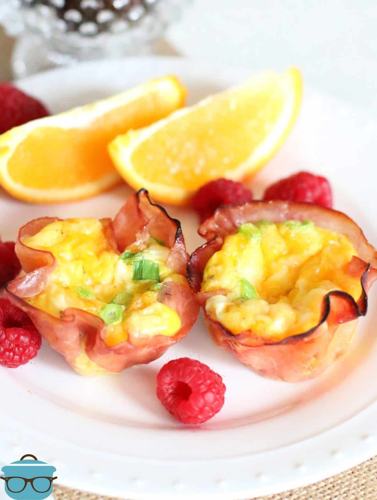 two ham and egg cups shown on a white plate with fresh raspberries and sliced oranges. 