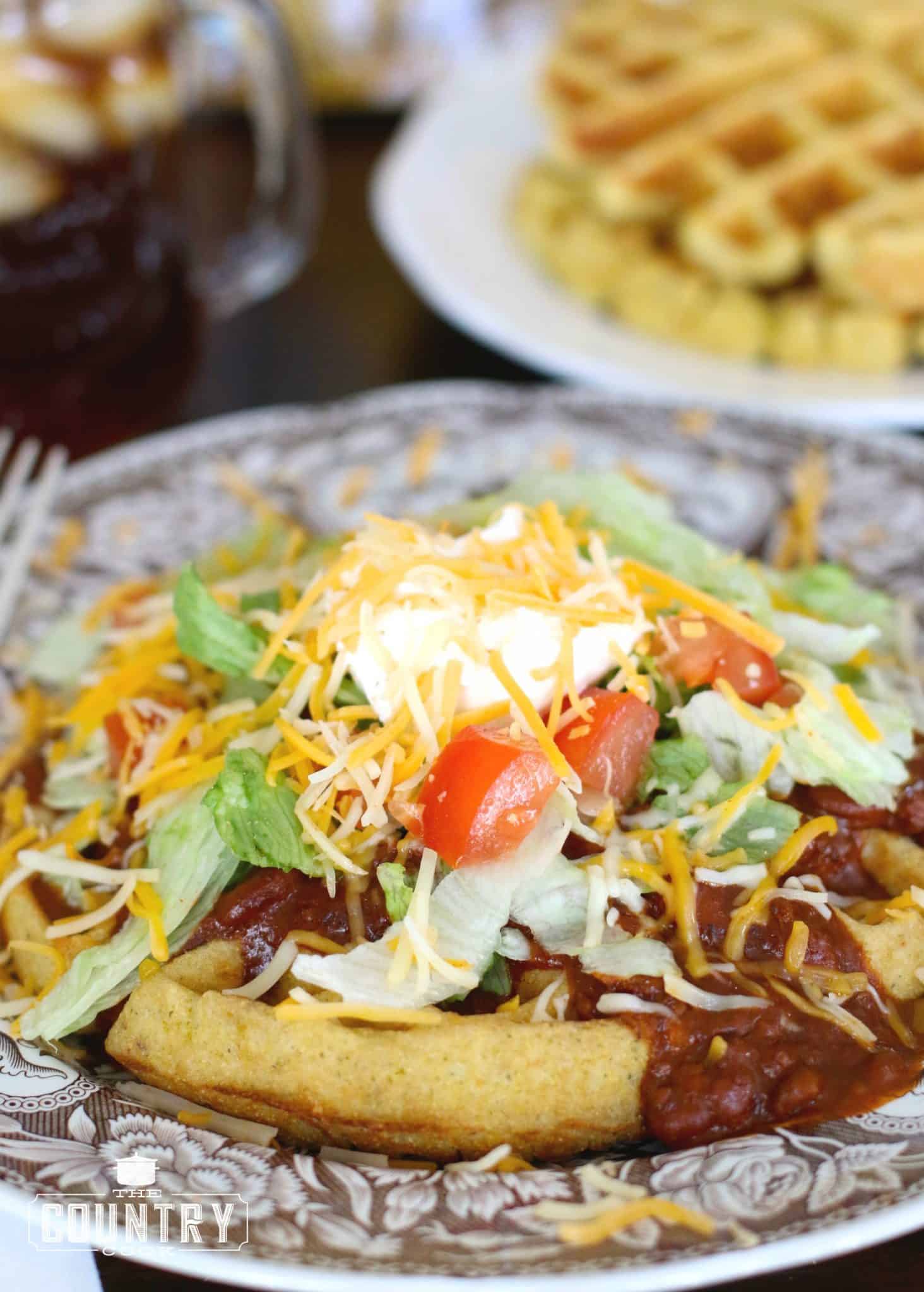 cornbread waffle shown on a brown and white plate. Waffle is topped with chili, lettuce, shredded cheese, sour cream and diced tomatoes. 