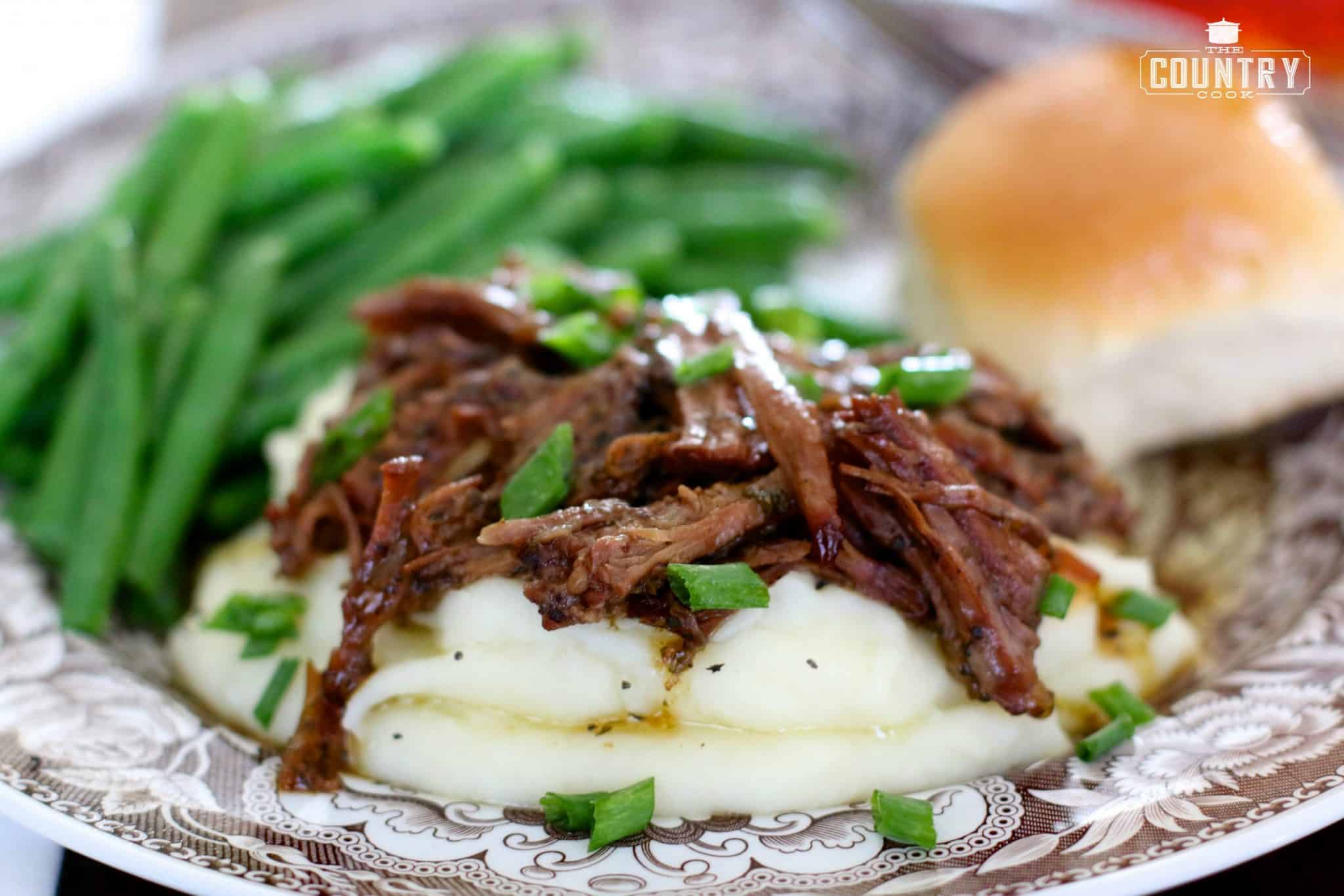 a serving of shredded pot roast on a pile of mashed potatoes with a side of green beans and a roll.