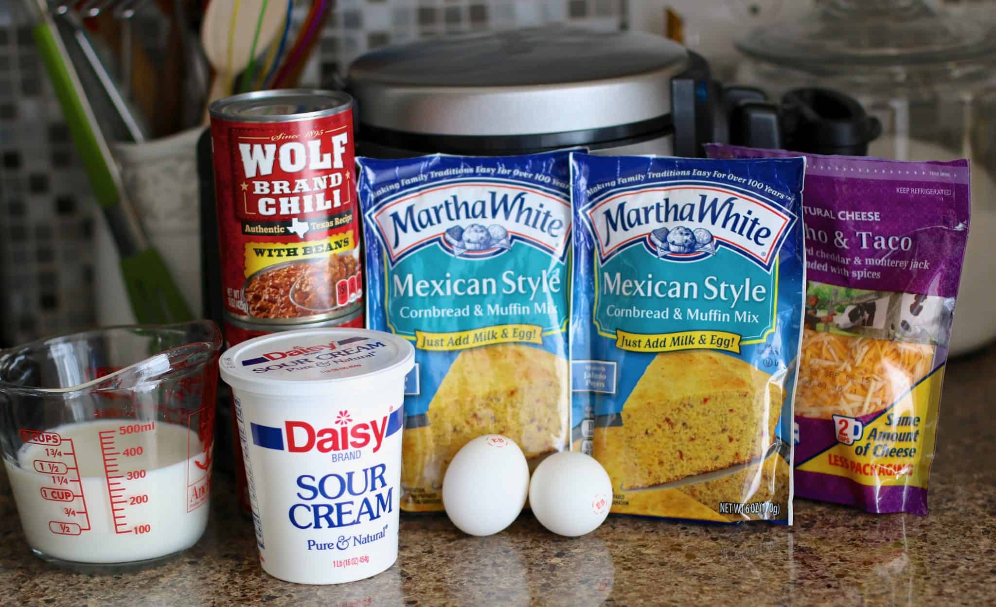 two packets of Mexican Style cornbread, two cans of wolf brand chili, shredded cheddar cheese, two eggs, a tub of sour cream and milk in am measuring cup. 